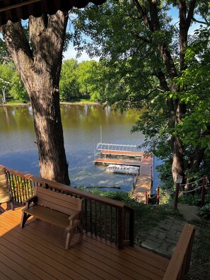Ein Holzdeck mit einer Bank mit Blick auf einen ruhigen See, eine Anlegestelle mit Booten und ein großer Baum.