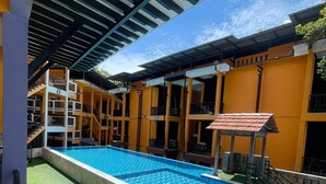A swimming pool with a tiled deck, a covered seating area, and a building with multiple balconies.