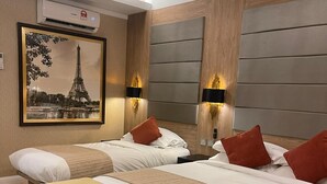 A hotel room with two beds, a bench, a framed picture of the Eiffel Tower, and wall-mounted lights.
