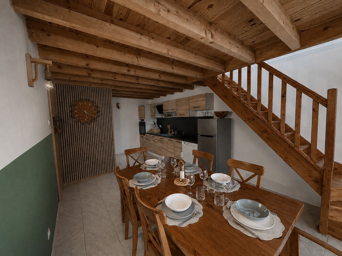 Un coin repas avec une table en bois dressée pour six personnes, une cuisine en arrière-plan et un escalier en bois.
