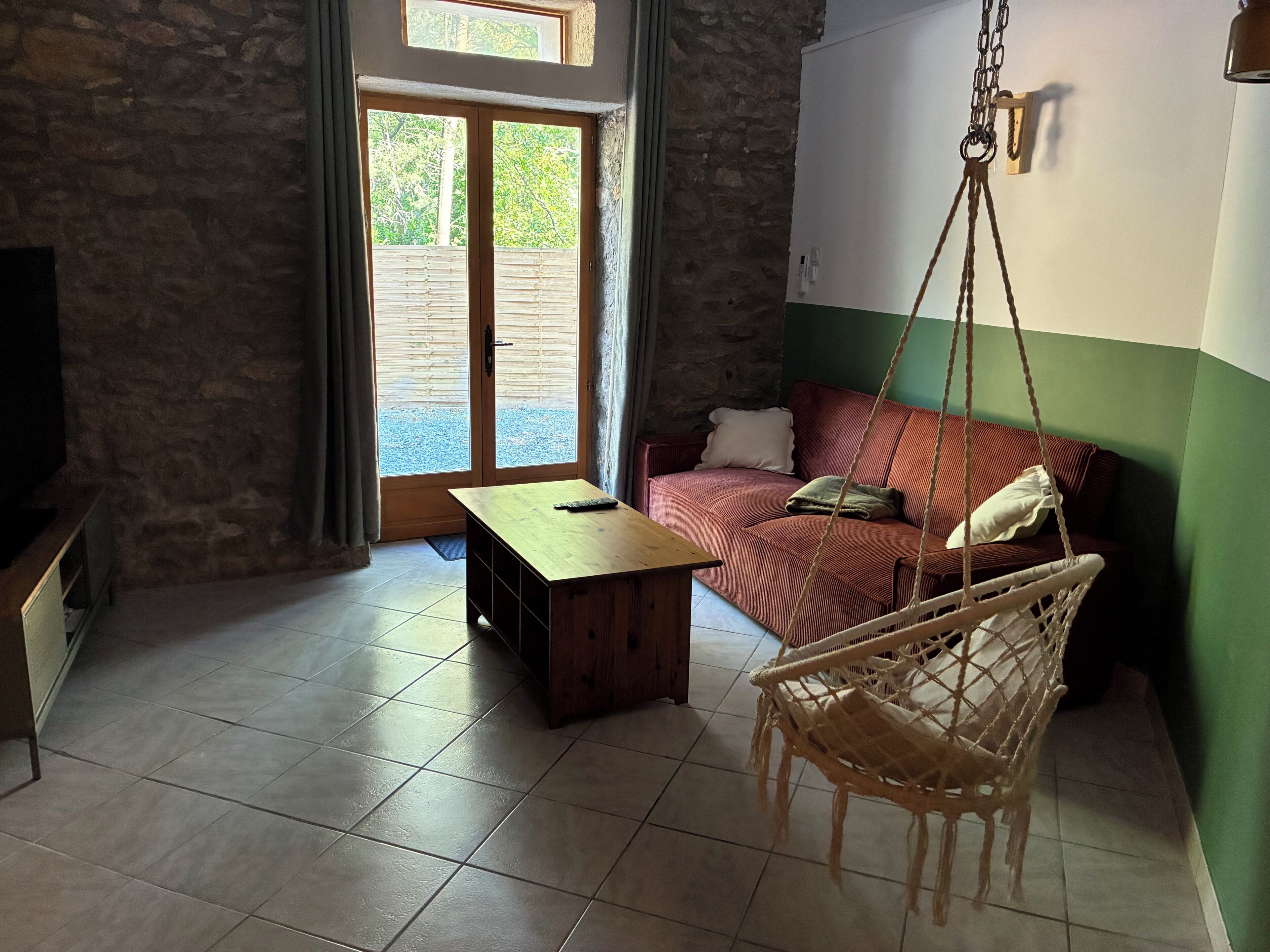A room with a sofa, a hanging chair, a wooden table, and a stone wall.