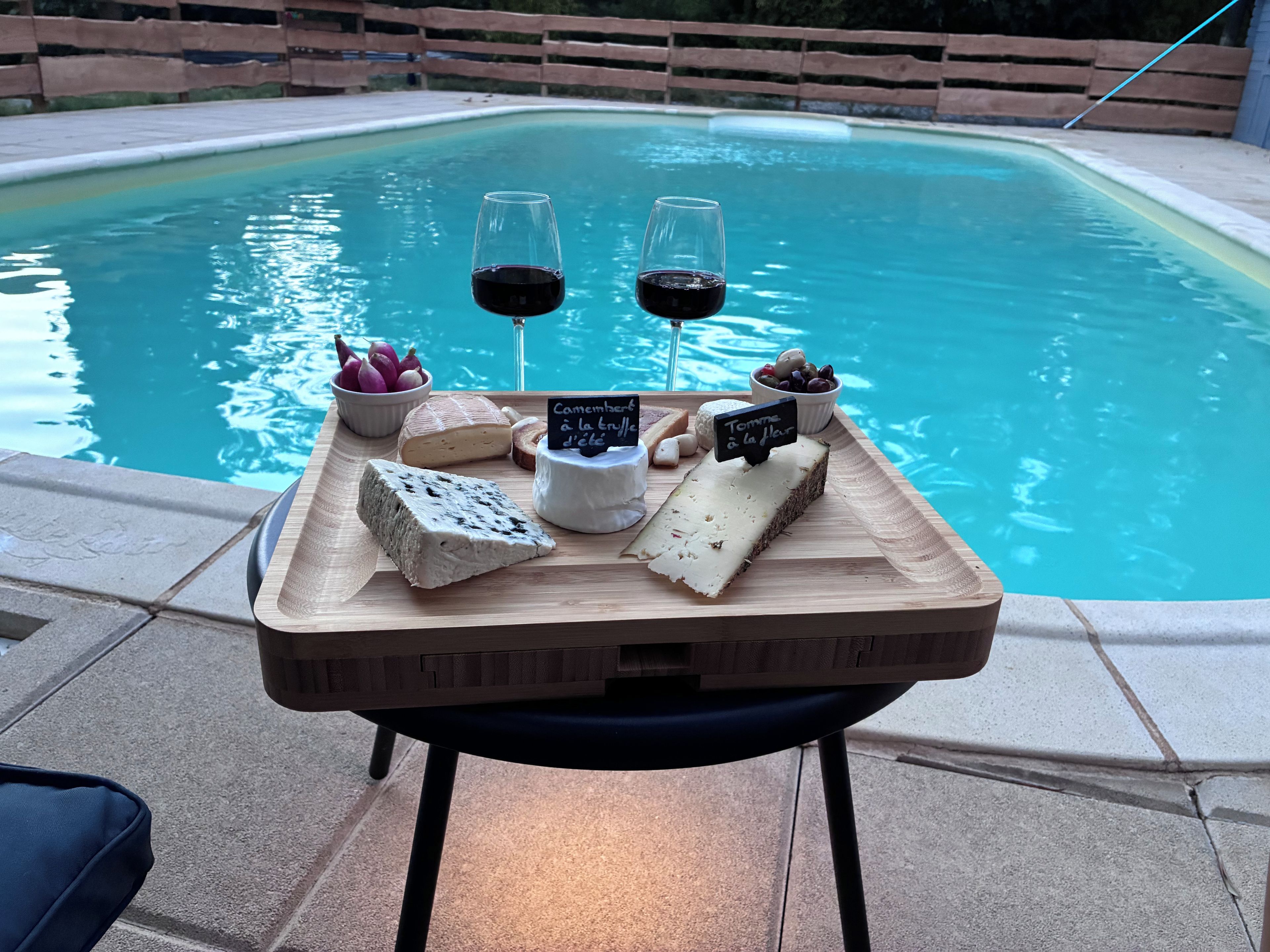 Mesa junto a la piscina con una tabla de quesos, dos copas de vino tinto y un cartel que indica variedades de queso Camembert y Tomme.