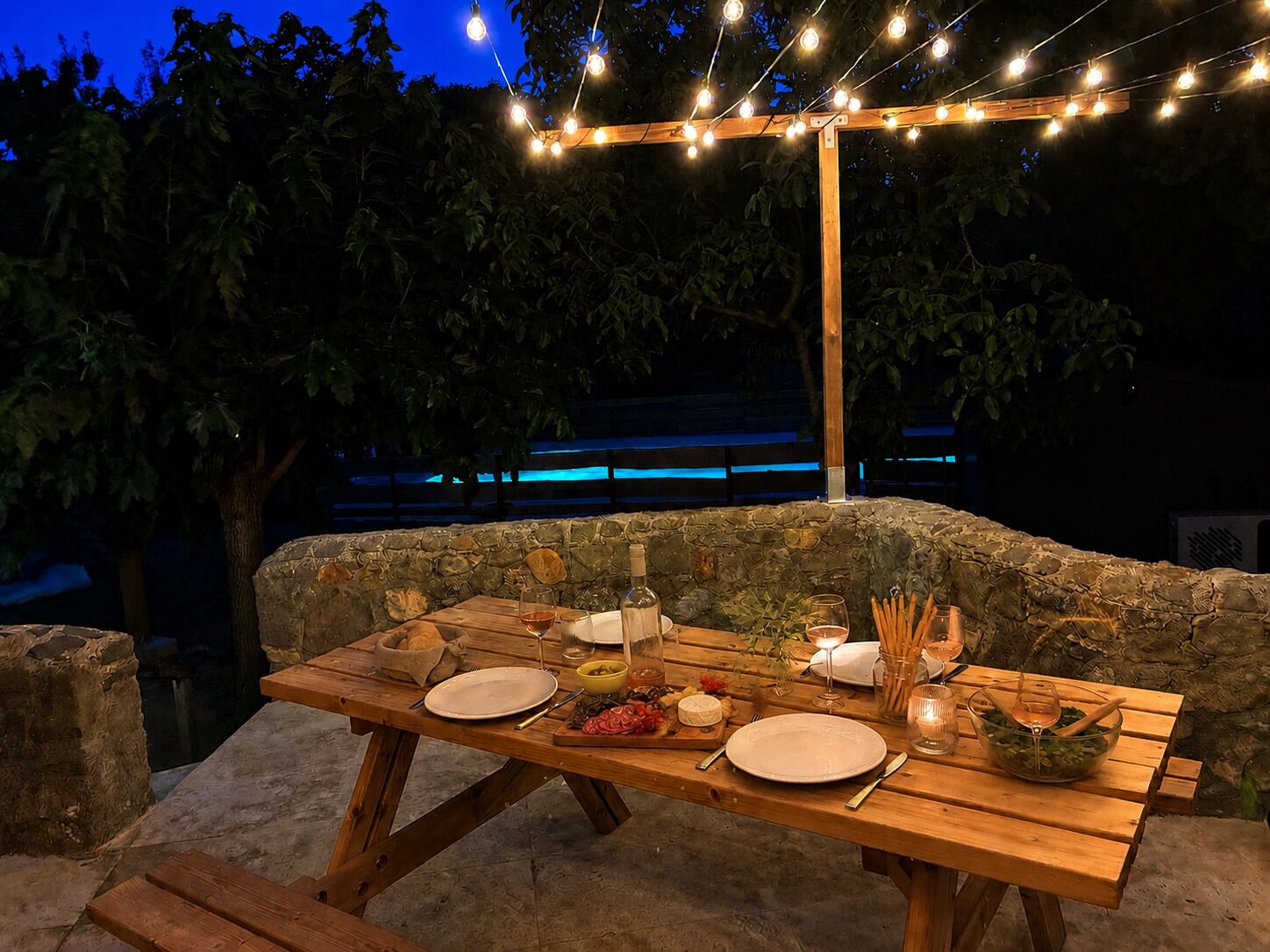 An outdoor dining area with a stone wall backdrop, wooden table, and string lights.