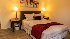 Un letto ben rifatto con una coperta rossa, due lampade ai lati e un quadro appeso sopra il letto, oltre a un cestino.