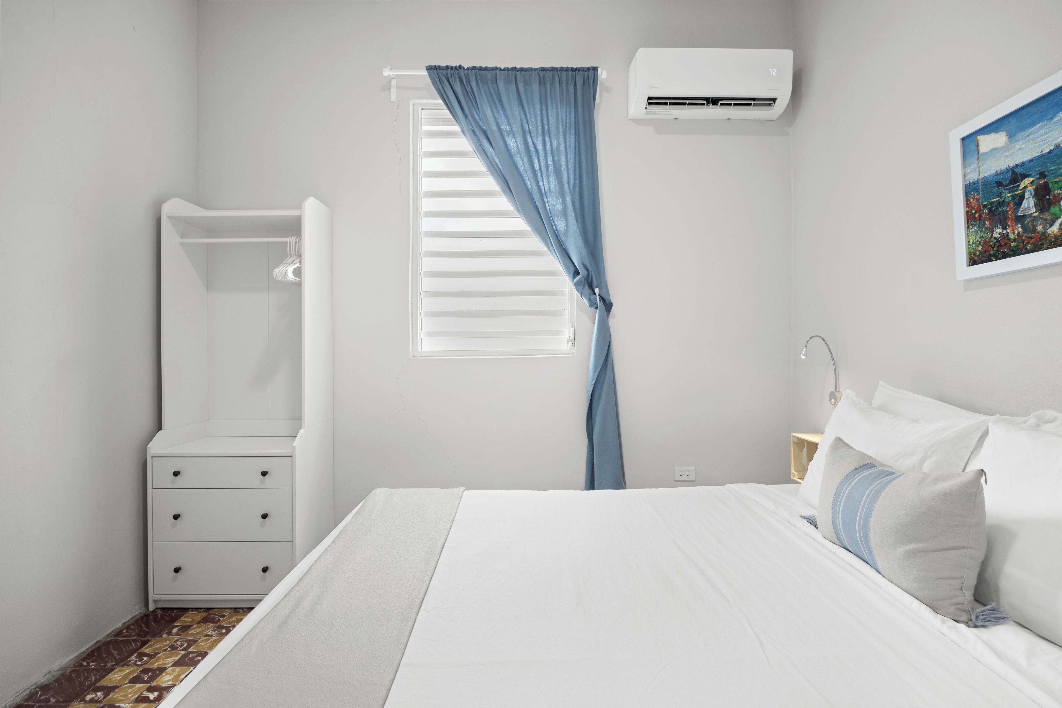 A bedroom with a bed, a nightstand, a dresser, a window with blinds, and an air conditioner.