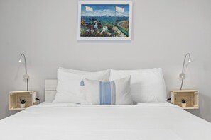 Ein ordentlich bezogenes Bett mit weißen Leinen, zwei Nachttische mit Lampen, ein an der Wand hängendes Bild und ein kleiner Topfpflanze auf einem der Tische.