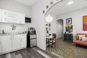 A modern kitchen with white cabinets, a built-in microwave, and a refrigerator. There is a dining area with a table and chairs, and a sofa in the living room.