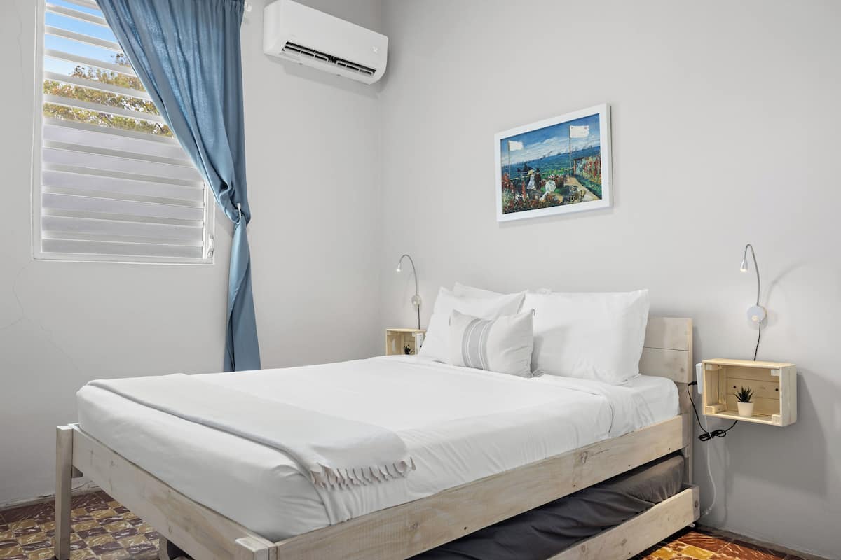 Ein Schlafzimmer mit einem Bett, einer Wand-Klimaanlage, einem Bild, Nachttischlampen und einem kleinen Regal.