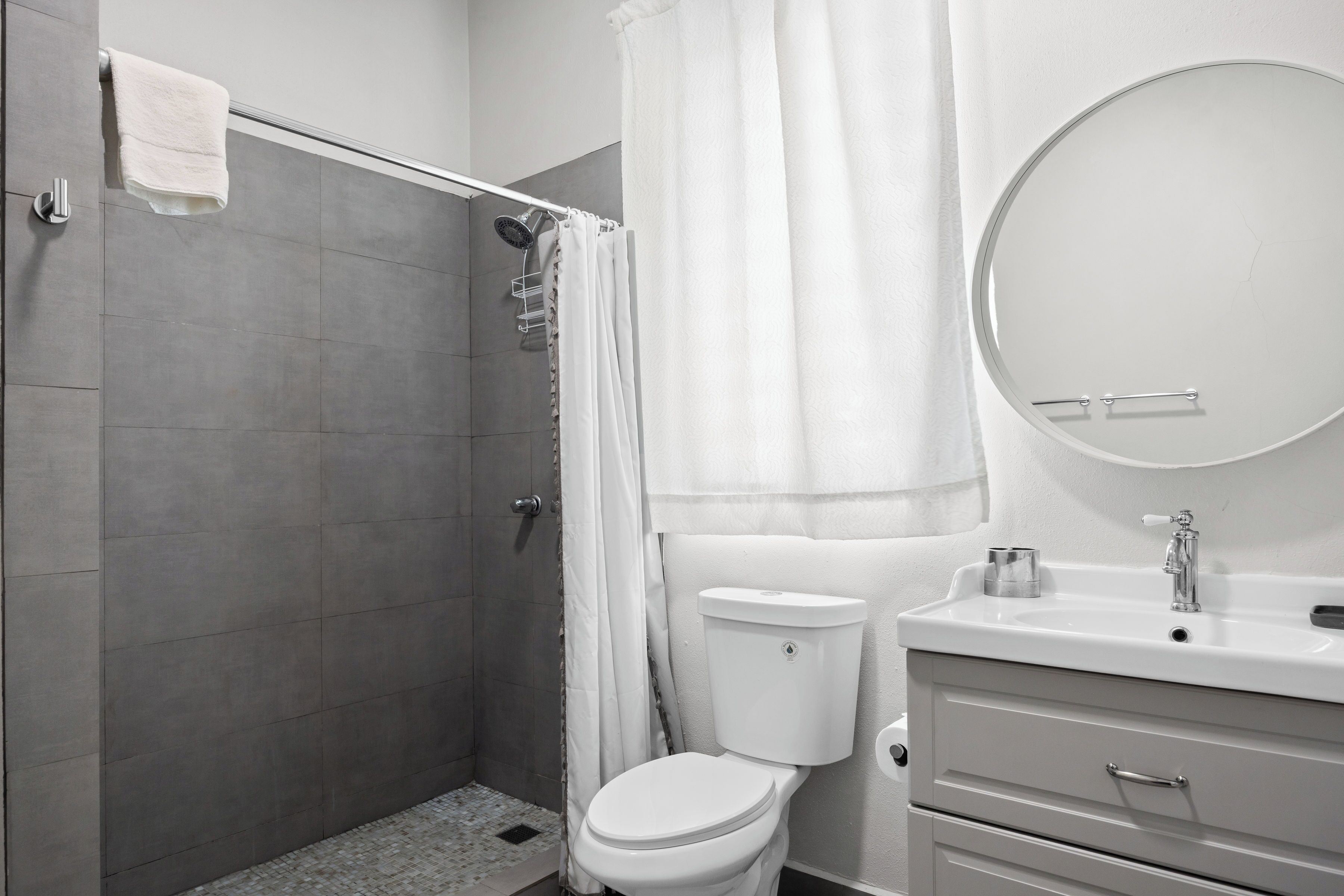 A bathroom with a shower, toilet, sink, and a round mirror.