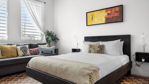 A bedroom with a bed, a sofa, a plant, a painting, and a nightstand.