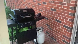 Una parrilla para barbacoa en una terraza de madera, un tanque de gas y un muro de ladrillo.