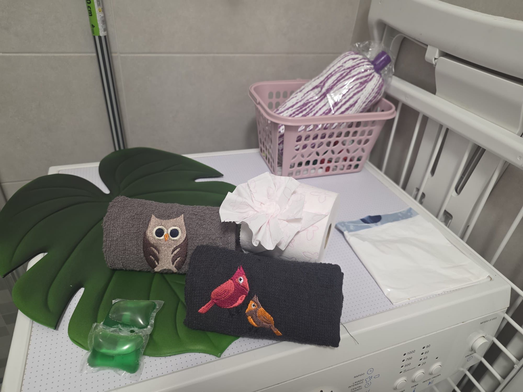 A crib with a white mattress, a pink basket with a striped item, a green leaf, and a cloth with bird embroidery.