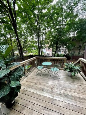 Eine Holzterrasse mit Tisch und Stühlen, umgeben von großen Grünpflanzen und Bäumen.