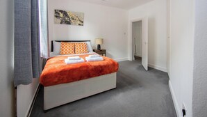 Une chambre d’hôtel avec un lit, une table de chevet, une lampe, un tableau au mur et une porte donnant sur une autre pièce.