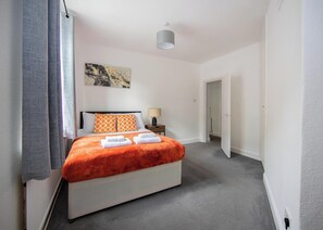 Ein Hotelzimmer mit einem Bett, einem Nachttisch, einer Lampe, einem Gemälde an der Wand und einer Tür zu einem anderen Raum.