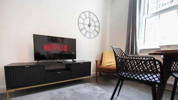 Un salon avec un téléviseur affichant le logo « NETFLIX », un meuble de rangement noir, un fauteuil à motifs et une horloge murale.