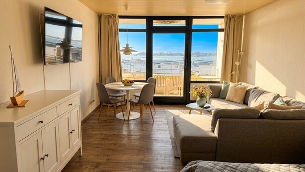 A room with a sofa, a dining table with chairs, a TV mounted on the wall, and a large window with a view of buildings. - Sea view in the Meerzeitliebe Fehmarn (Fehmarn)