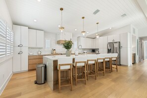 A modern kitchen with white cabinetry, wooden bar stools, and pendant lighting. - New AMI Retreat | Private Pool | Walk to Beach (Anna Maria)