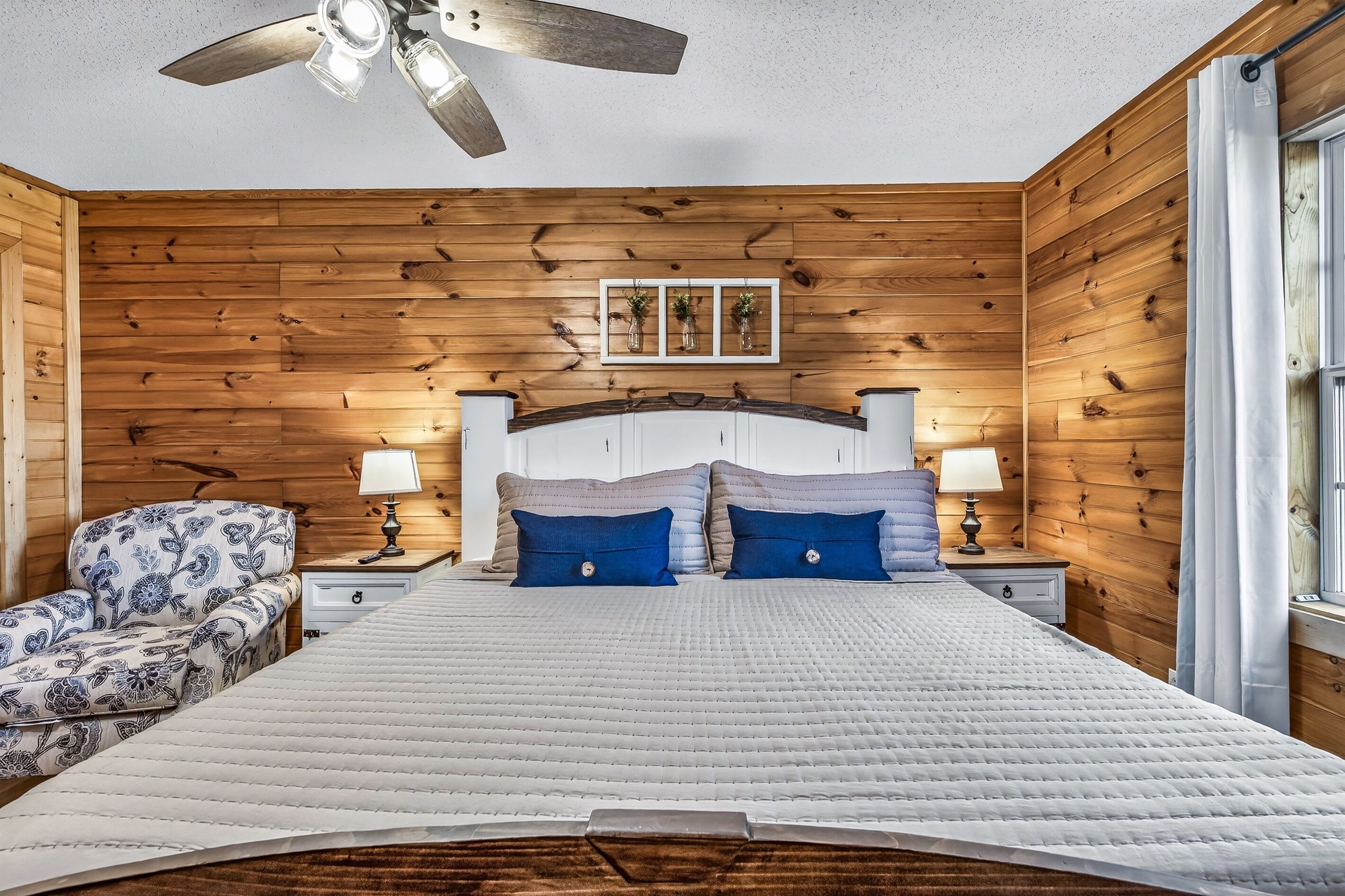 Une chambre avec des murs lambrés en bois, un lit avec une literie blanche et des oreillers bleus, une table de chevet avec une lampe, une chaise à motifs floraux et un ventilateur de plafond.