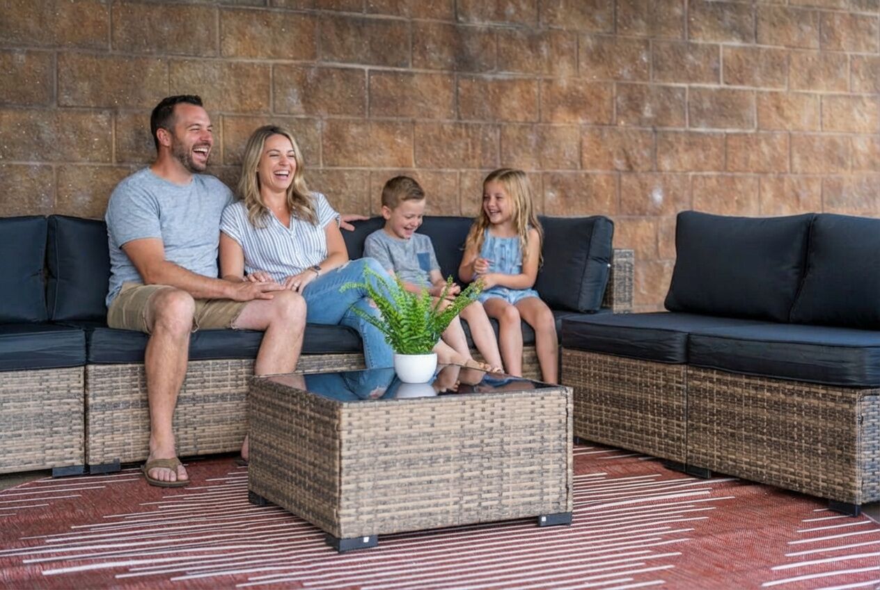 A family on a dark-colored sofa with a woven coffee table in front, set against a stone wall.
