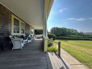 A covered porch with outdoor furniture, a view of a green lawn, and a building with a yellow facade. - North Chatham Beachwalk -Spacious 6 bedroom walk to beach sprawling private yard (Chatham)
