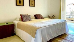 A bedroom with a bed, a nightstand, a chair, and a view of the outside. - Mother Africa Apartment – ​​5 minutes from Primeira Praia (First Beach) (Morro de São Paulo)