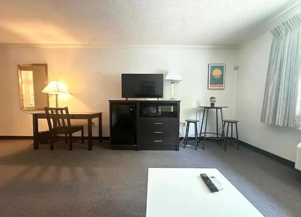 A hotel room with a TV, a desk, a chair, a lamp, a small table, and a remote control on a white table. - Kiteville Phoenixville (Phoenixville)