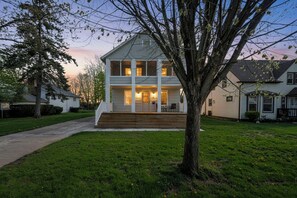 A two-story house with a front porch, a large tree in the yard, and a driveway leading to the house. - Bright 2BR w/ Sunroom Near Downtown & Beach (Euclid)