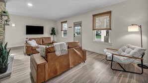 A living room with a brown leather sofa, a wooden floor, a standing lamp, and a television mounted on the wall. - Bright 2BR w/ Sunroom Near Downtown & Beach (Euclid)