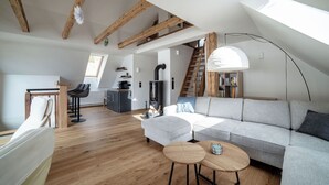 Un loft moderne avec des poutres apparentes, un lit en mezzanine, un canapé gris, une table basse ronde et un escalier menant à un niveau supérieur.