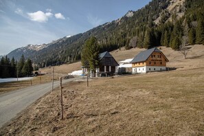 Un paisaje rural con una casa de madera de dos pisos, una carretera y montañas al fondo.