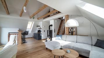 Ein großzügiges Loft mit hoher Decke, freiliegenden Holzbalken, einer modernen Küche, einer Sitzecke mit einer Couch und zwei kleinen Tischen sowie einer Treppe, die zu einer oberen Ebene führt.