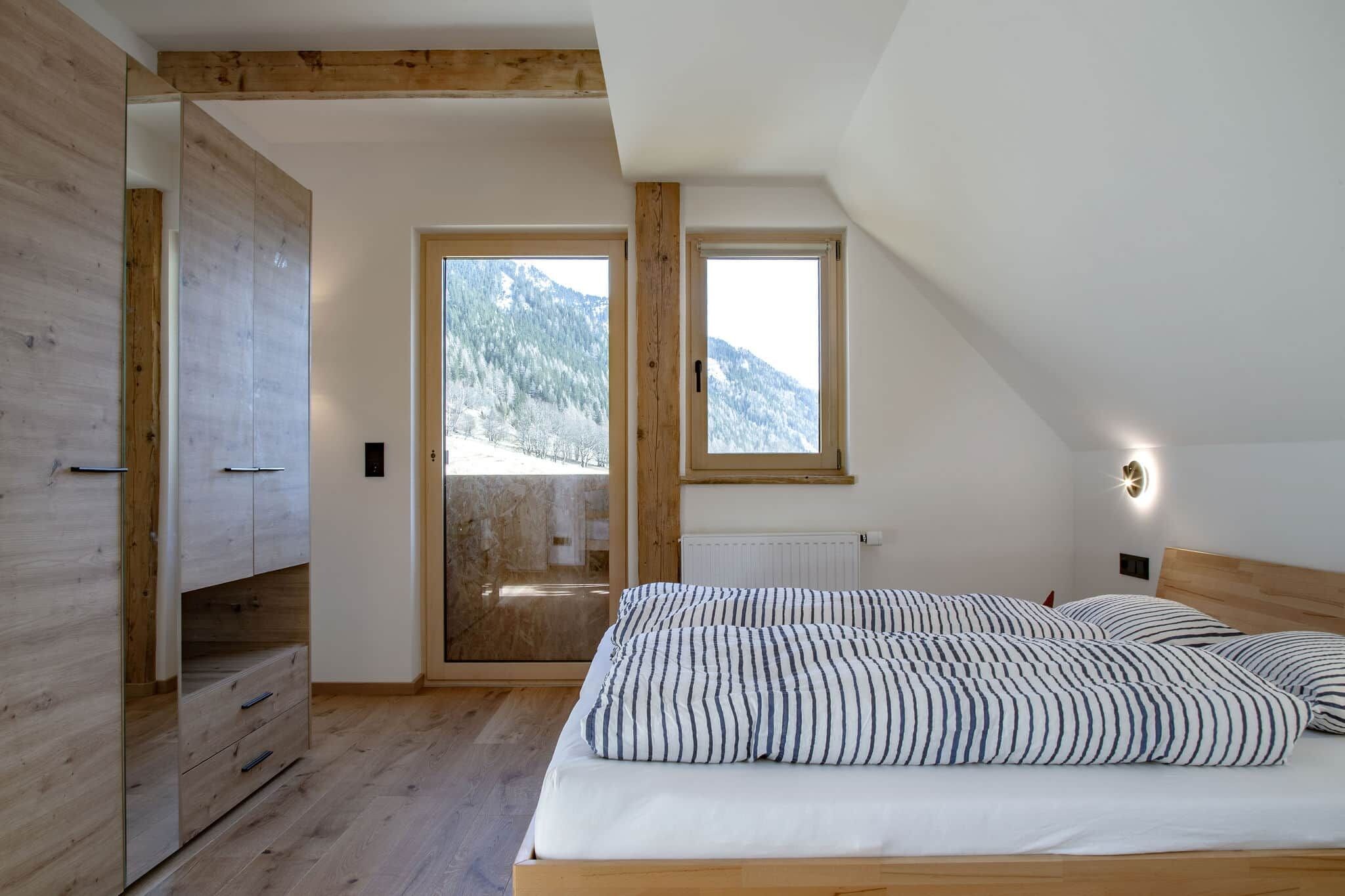 Un dormitorio con techo de madera, una cama con ropa de cama a rayas, una ventana que ofrece vistas a montañas nevadas y un radiador.