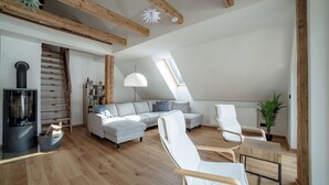 Un loft moderne avec des poutres apparentes, une cheminée intégrée, un canapé gris, des fauteuils blancs et une lucarne.