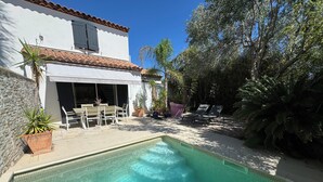Una piscina con solárium empedrado, un patio con mesa y sillas, y un edificio con tejado y ventanas de cerámica.