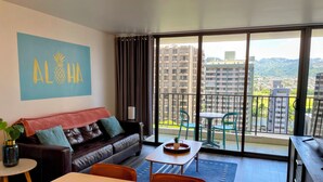 A modern living room with a sofa, a dining table, and a view of a cityscape through large windows. - Waikiki Banyan Pineapple Suite with AC & Free Parking (Honolulu)