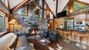 A cozy living room with a stone fireplace, leather seating, and a kitchen with modern appliances. - Grand Elk Chalet by Avantstay Private Beach + Pier + Hot Tub Historic A-frame in Carnelian Bay Near Tahoe City + Hiking Trails (Carnelian Bay)