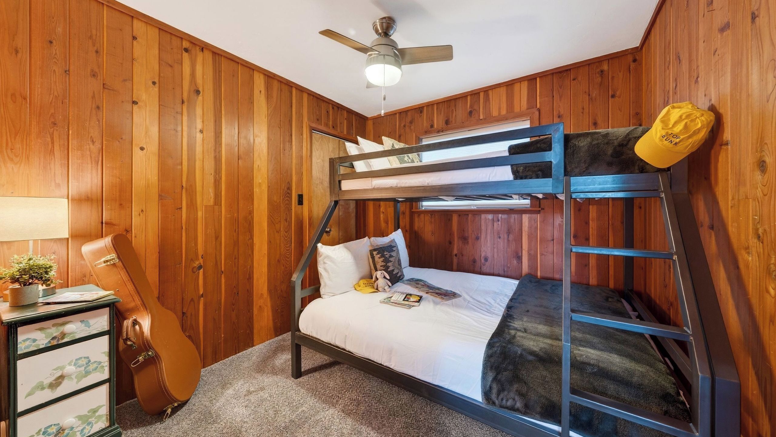 A bunk bed room with wooden walls, a ceiling fan, a desk with a lamp, and a suitcase.