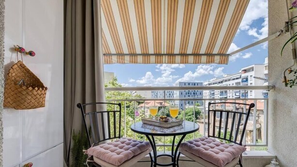 Ein Balkon mit zwei gepolsterten Stühlen, einem kleinen Tisch mit Getränken, einem gewebten Korb und einem Blumentopf.