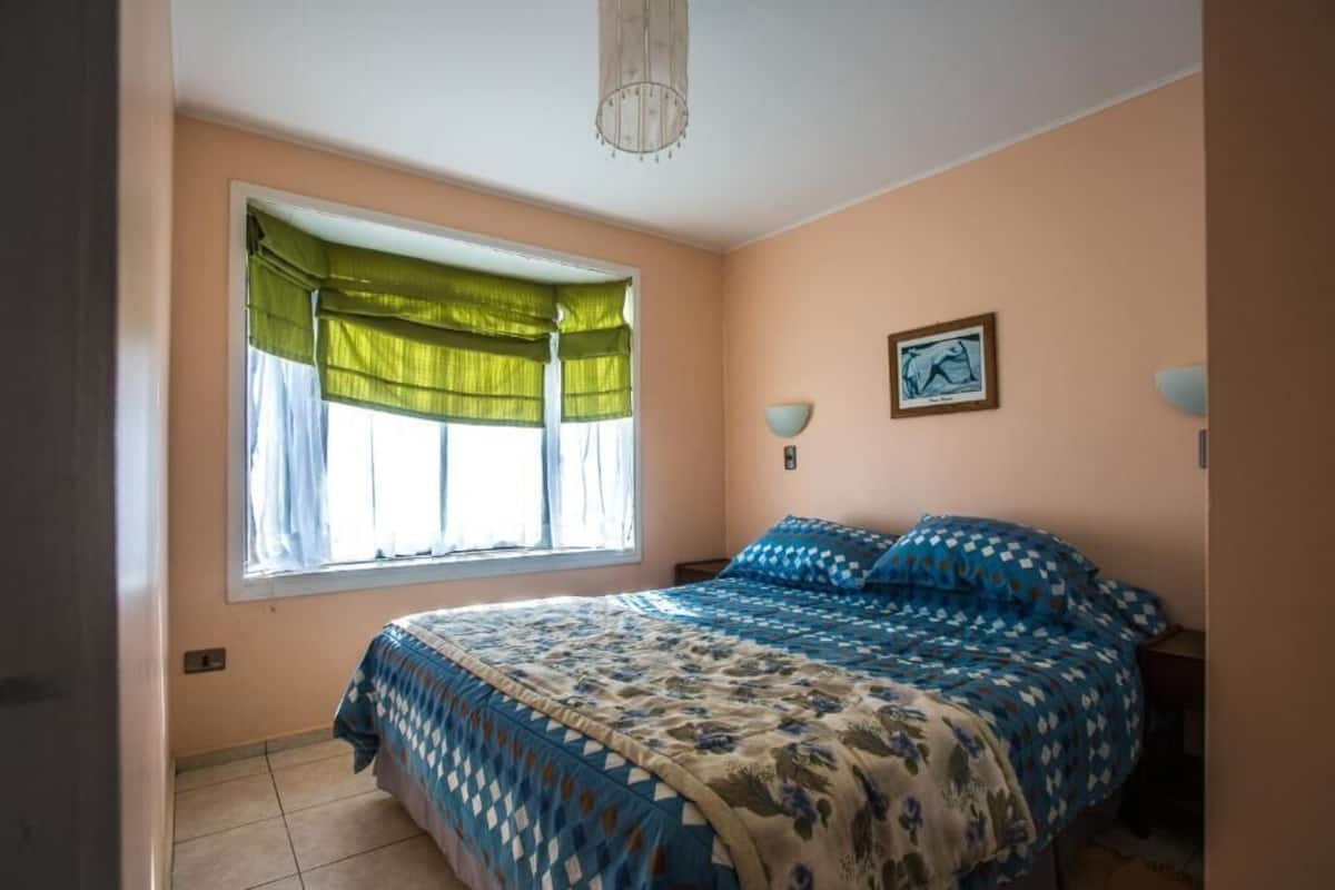 A bedroom with a bed, a window with curtains, a wall-mounted lamp, and a framed picture.
