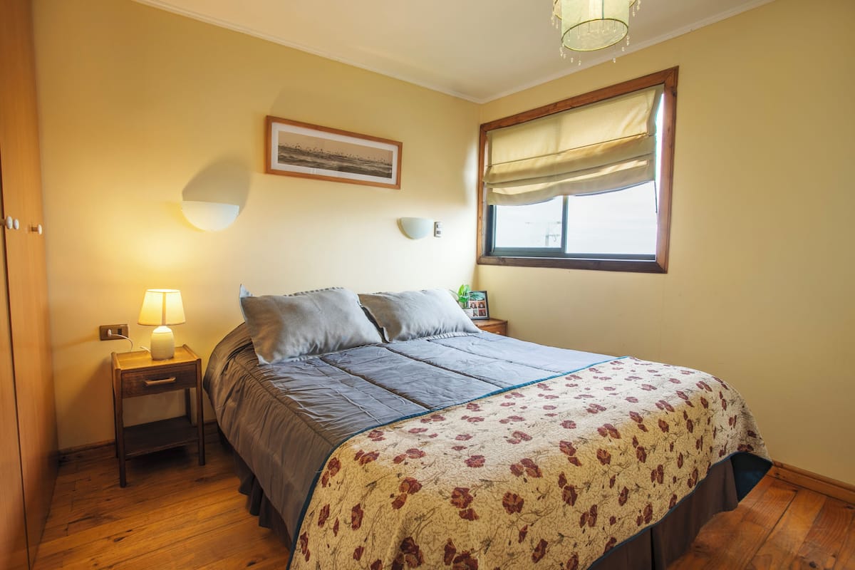 A bedroom with a bed, bedside lamp, nightstand, window with curtains, wall-mounted light, and a framed picture.