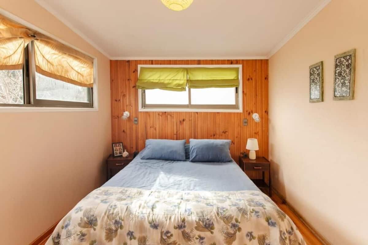 A bedroom with a wooden headboard, a bed with floral bedding, two bedside tables with lamps, and a window with a green valance.