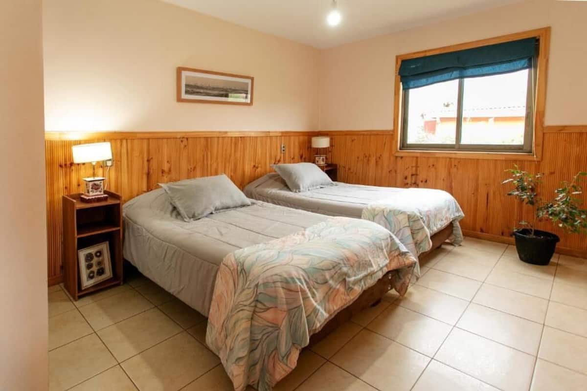 A room with two beds, wooden paneling, a window with a view, a bedside table with a lamp, and a potted plant.