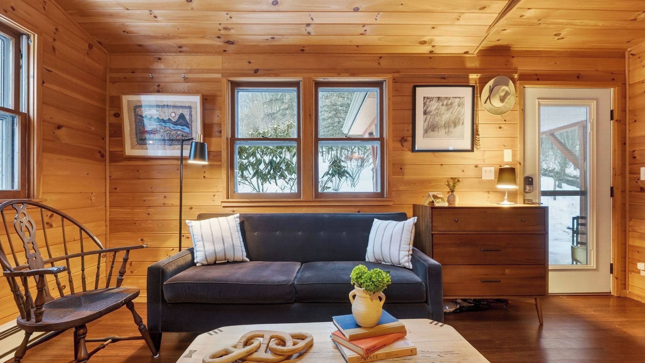 A cozy cabin interior with a dark sofa, wooden walls, a dining table with books, and a chair.