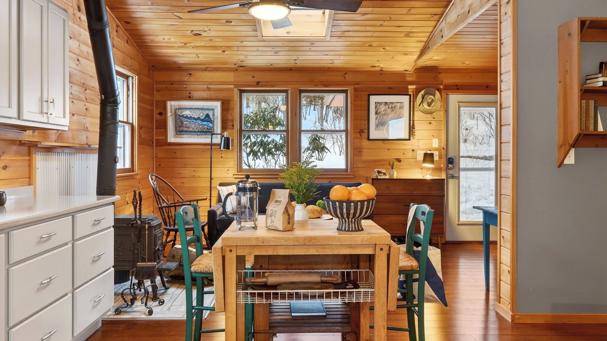 A wooden cabin interior with a dining table, chairs, a coffee maker, a rolling pin, and a window with a view of trees.