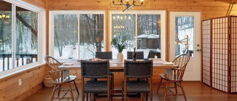 A wooden interior with a dining table, chairs, and large windows.