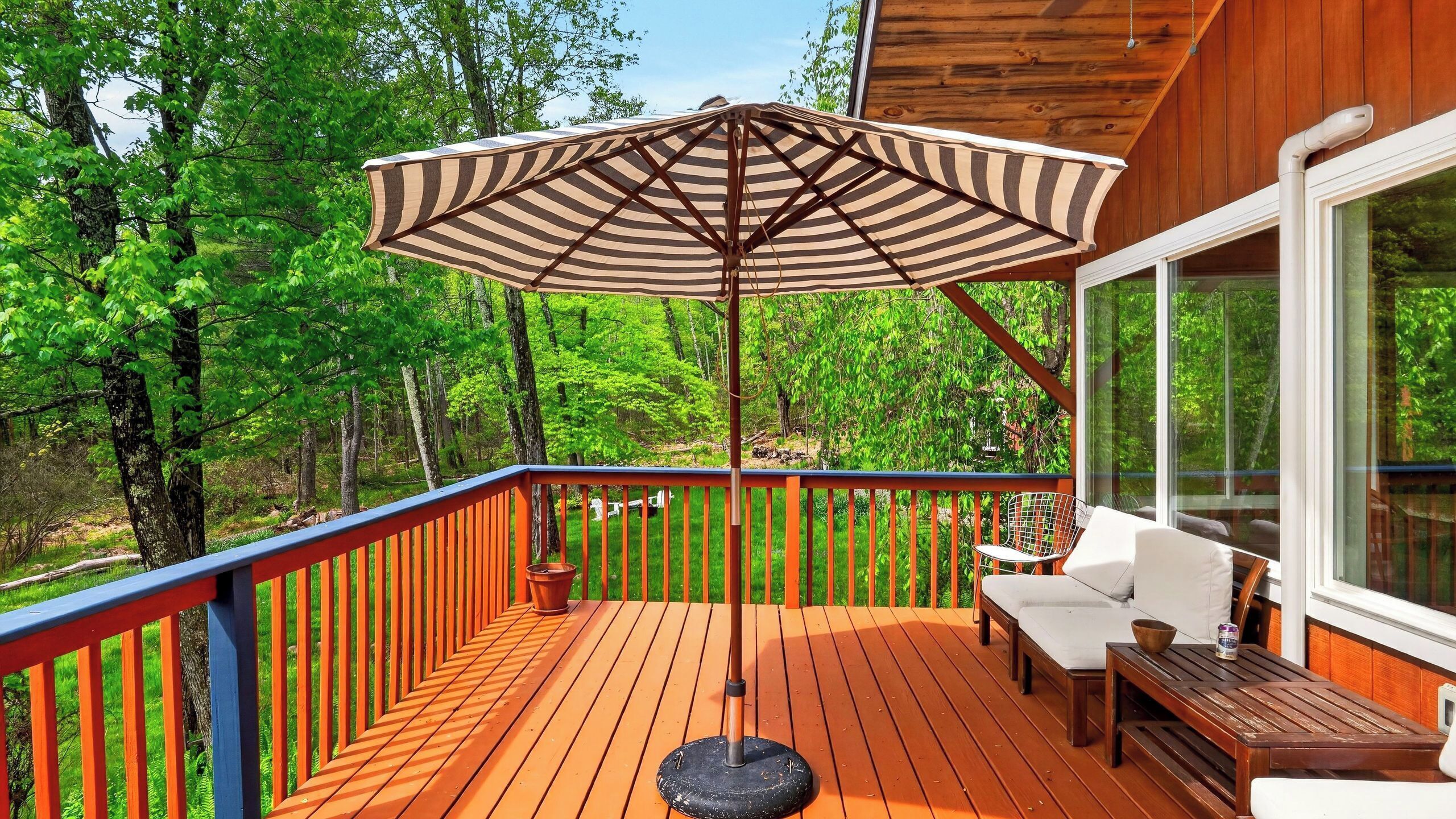 A wooden deck with a striped umbrella, outdoor seating, and a view of a forest.