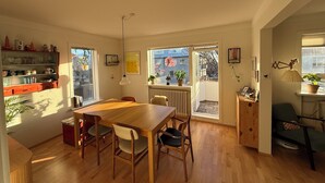 Ein Essbereich mit Holztisch und -stühlen, eine Küche mit Regalen und einem Heizkörper sowie ein Blick durch ein Fenster.