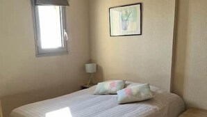 Quarto com cama, mesa de cabeceira, janela com cortinas, luminária na parede e um quadro emoldurado.