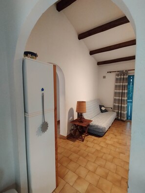 Ein Zimmer mit Fliesenboden, einem Bett mit gemustertem Überwurf, einem kleinen Tisch mit Lampe und einer weißen Tür mit dekorativer Griffleiste.